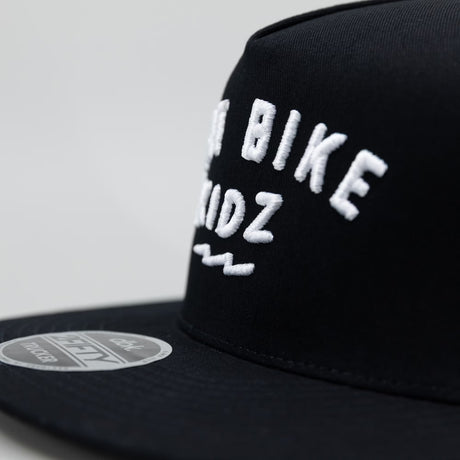 Black adult 12 O'Clock Trucker Hat close-up showing white embroidered Dirt Bike Kidz arch logo detail on structured front panel