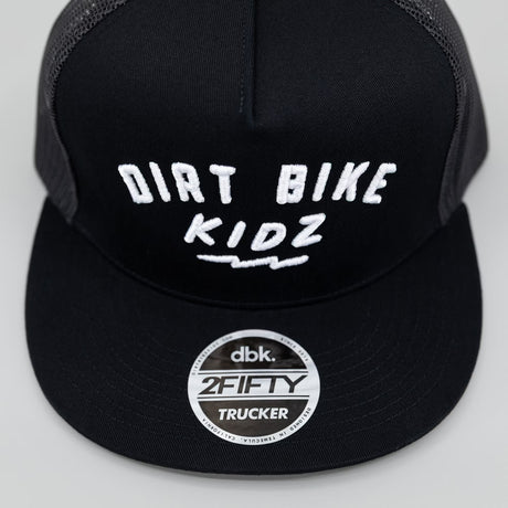 Black adult 12 O'Clock Trucker Hat close-up of brim showing DBK 2Fifty Trucker sticker and white embroidered Dirt Bike Kidz arch logo detail