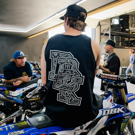 Black adult tank top back view with oversized detailed bandana-style DBK graphic, worn by man sitting on a DBK Yamaha dirt bike surrounded by riders in a motocross garage setting