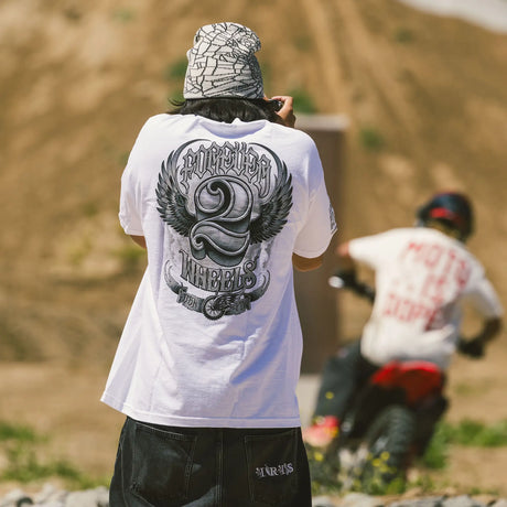 Person wearing a white Forever 2 Wheels t-shirt with a graphic design, standing on a dirt track with another person and a motorcycle in the background.
