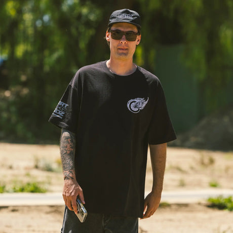 Man wearing a black Forever 2 Wheels t-shirt with a logo, sunglasses, and a cap outdoors.