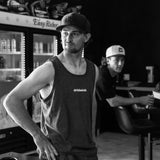 Dark charcoal adult tank top front view with white Dirt Bike Kidz chest logo, worn by man in a DBK snapback hat in a lifestyle setting