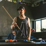 Dark charcoal Moto is Dope adult tank top front view with white Dirt Bike Kidz chest logo, worn by man holding a pool cue in a moto-themed game room with helmets displayed in the background