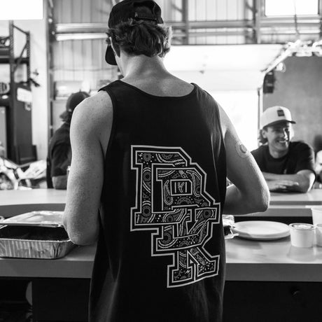 Black adult tank top back view with oversized detailed bandana-style DBK graphic, worn by man in a casual garage hangout setting