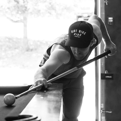 Black adult 12 O'Clock Trucker Hat with white embroidered Dirt Bike Kidz arch logo, worn by man leaning over a pool table taking a shot