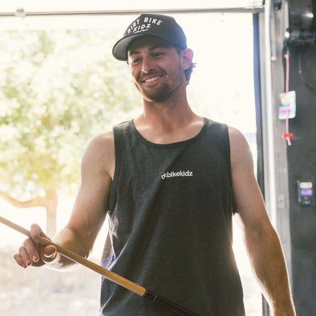 Black adult 12 O'Clock Trucker Hat with white embroidered Dirt Bike Kidz arch logo, worn by smiling man holding a pool cue in an outdoor garage setting, paired with dark charcoal Dirt Bike Kidz tank top