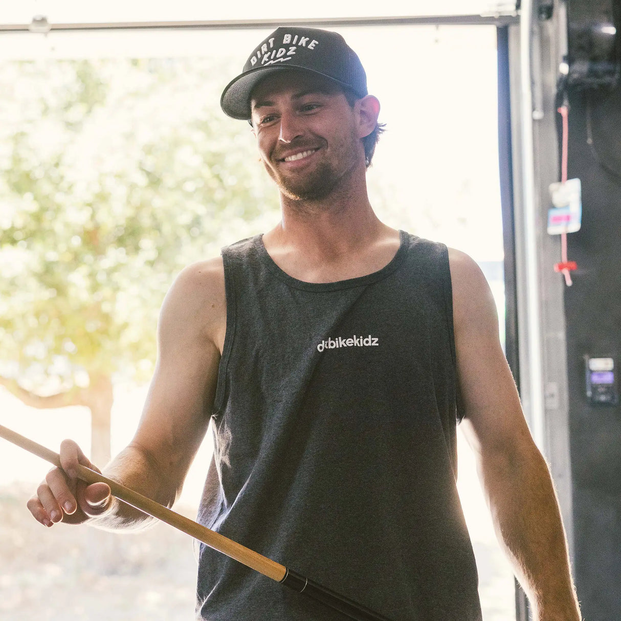Dark charcoal adult tank top front view with white Dirt Bike Kidz chest logo, worn by smiling man in a Dirt Bike Kidz snapback hat in an outdoor garage setting