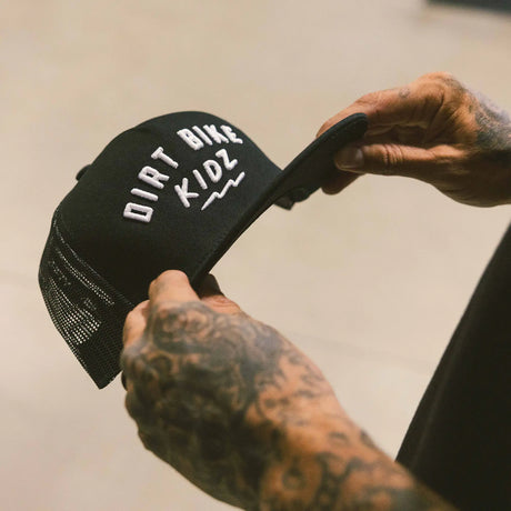 Black adult 12 O'Clock Trucker Hat with white embroidered Dirt Bike Kidz arch logo and black mesh back, held by tattooed hands showing embroidery detail