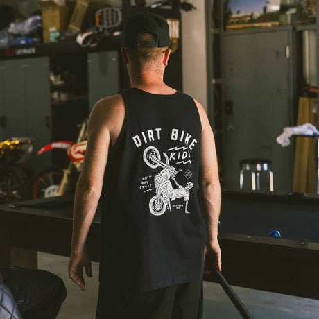 Black adult 12 O'Clock Tank Top back view with white skeleton rider wheelie graphic, worn by man standing in a moto garage with dirt bikes and a pool table in the background