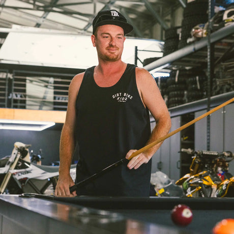 Black adult 12 O'Clock Tank Top front view with white Dirt Bike Kidz arch chest logo, worn by man holding a pool cue in a moto garage with dirt bikes in the background
