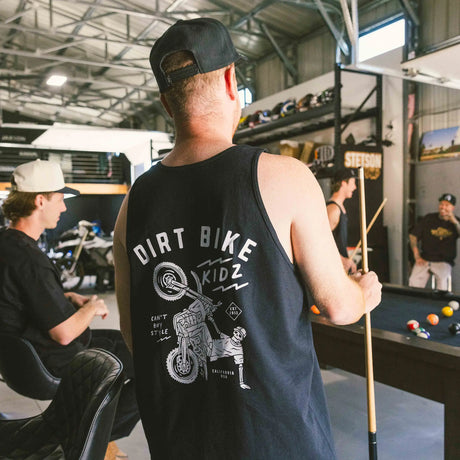 Black adult 12 O'Clock Tank Top back view with skeleton rider wheelie graphic, worn by man holding a pool cue in a moto garage hangout setting
