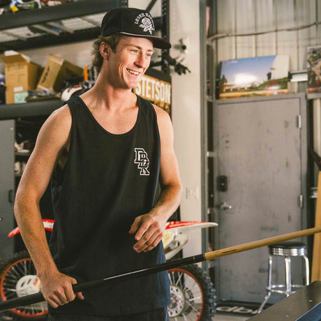 Black adult tank top front view with white Dropout chest logo, worn by smiling man holding a pool cue in a garage with dirt bikes in the background