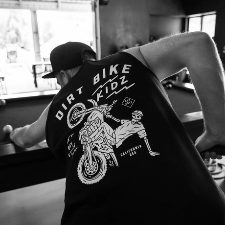 `Black adult 12 O'Clock Tank Top back view with skeleton rider wheelie graphic, worn by man leaning over a pool table in a garage setting`