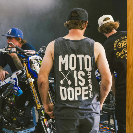 Dark charcoal adult tank top back view with bold "Moto Is Dope" graphic and Dirt Bike Kidz branding, worn at a motocross event with dirt bikes and riders in the background