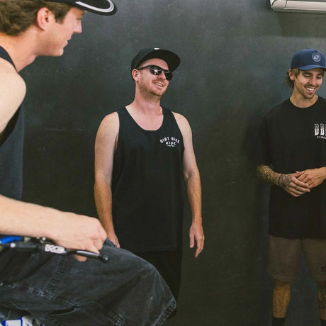 Black adult 12 O'Clock Tank Top front view with white Dirt Bike Kidz arch chest logo, worn by smiling man in a group of riders in a casual moto hangout setting