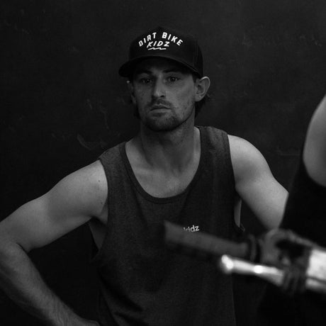 Black adult 12 O'Clock Trucker Hat with white embroidered Dirt Bike Kidz arch logo, worn by man on a dirt bike in a dark garage setting